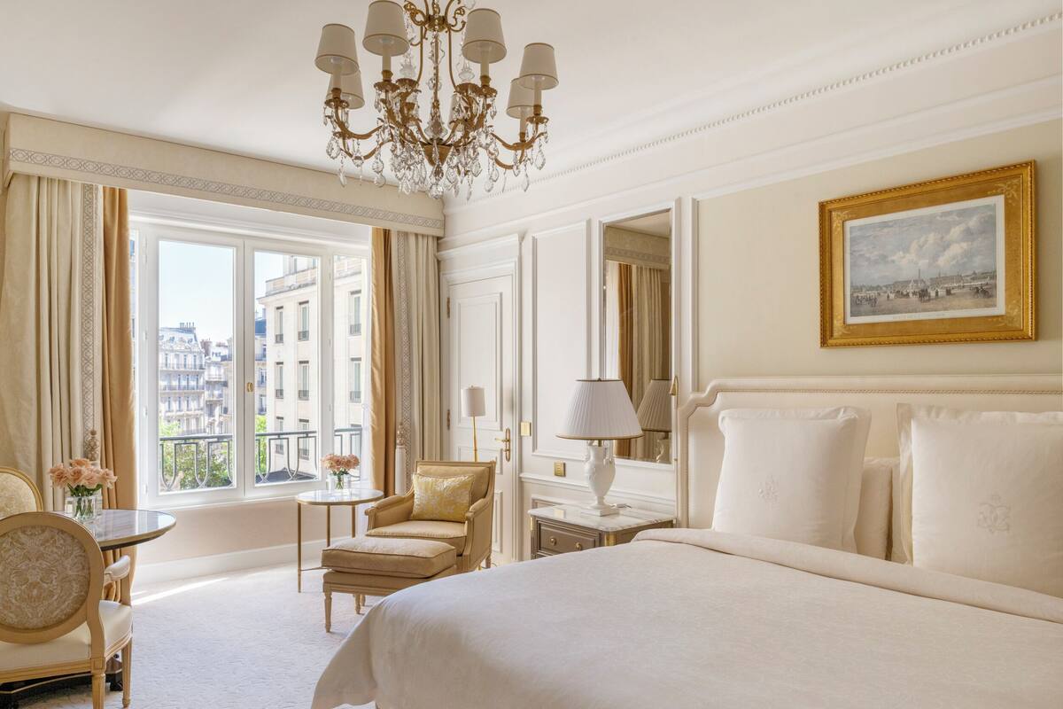 Une chambre d’hôtel dotée d’un grand lit, d’un coin salon avec un fauteuil et une table, d’un lustre et offrant une vue sur la ville par les fenêtres.