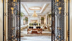 Un lobby de hotel opulento con una gran lámpara de araña, puertas de hierro negro ornamentadas y una mesa central decorada con arreglos florales.