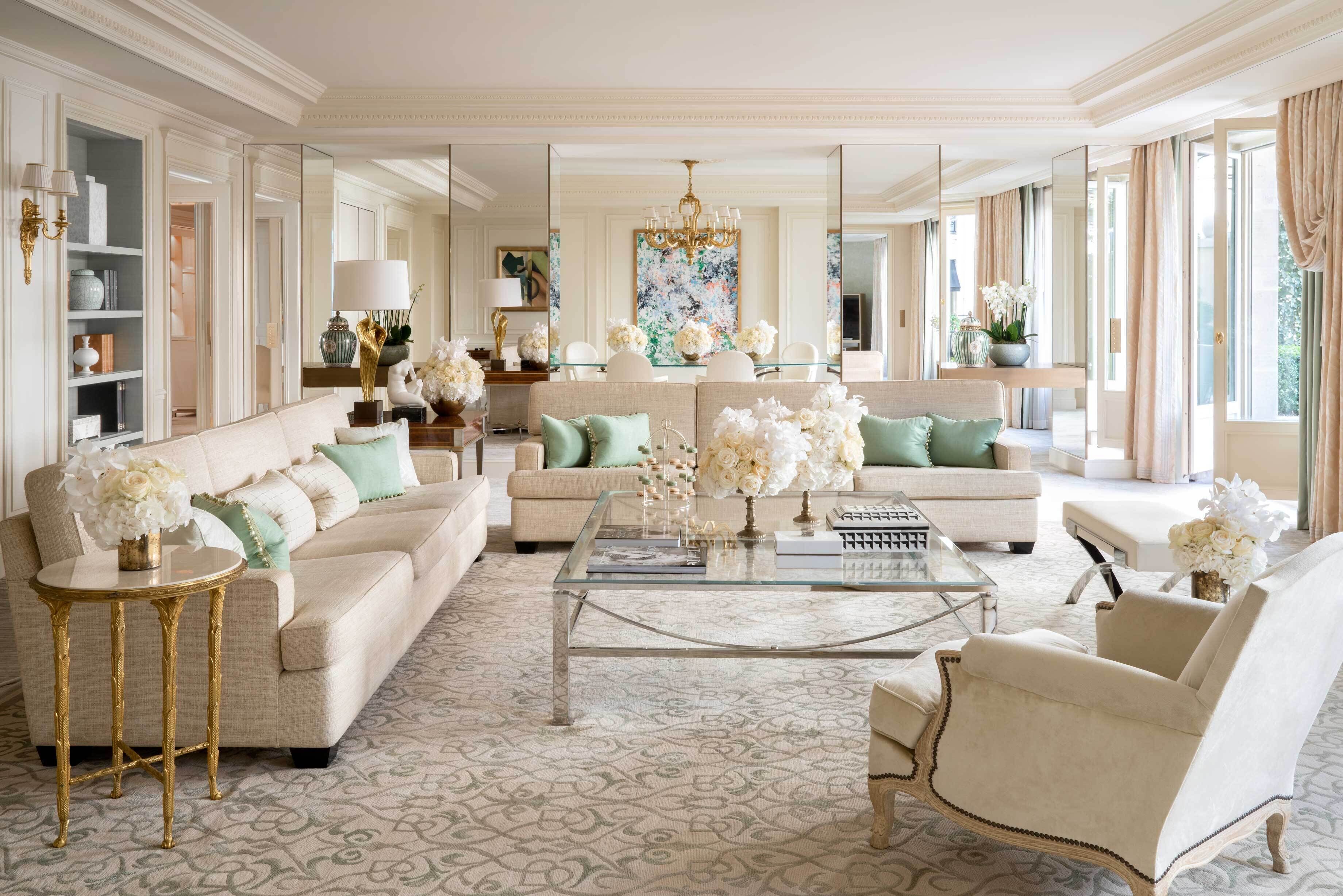 A spacious living room with a large sofa, a coffee table, and a chandelier. There are multiple seating options and a large mirror.