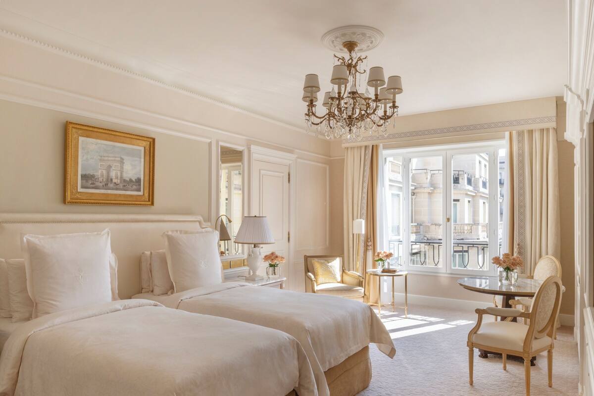 Une chambre d’hôtel avec deux grands lits, un lustre, un coin repas avec une table et des chaises, et une grande fenêtre avec des rideaux.