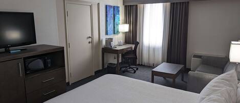 A hotel room with a bed, a desk with a chair, a television, and a window with curtains.