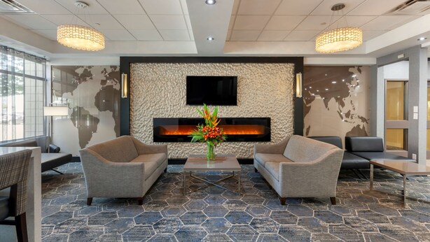A lobby with two sofas facing a wall-mounted TV and a fireplace. There are two large pendant lights and a coffee table with a vase of flowers. The floor has a geometric patterned tile design. - Best Western Plus Toronto Airport Hotel (Mississauga)