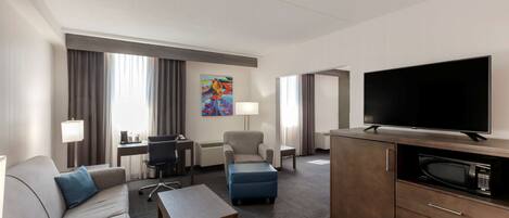 A hotel room with a sofa, ottoman, desk, chair, television, and a microwave.