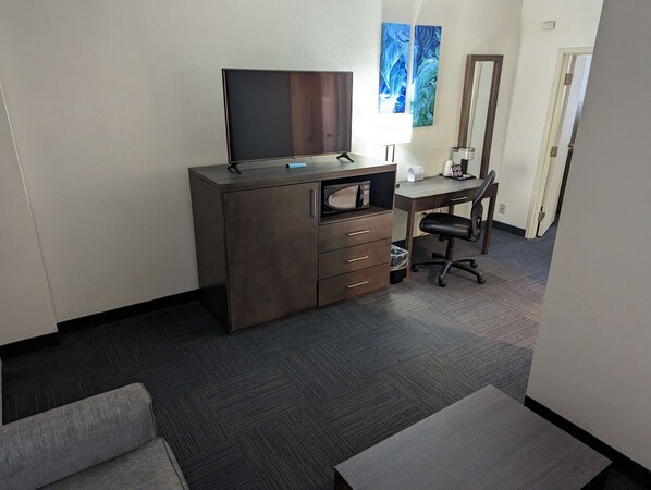 A hotel room with a dark wood dresser, a flat-screen TV, a desk with a chair, and a large window with blue artwork.