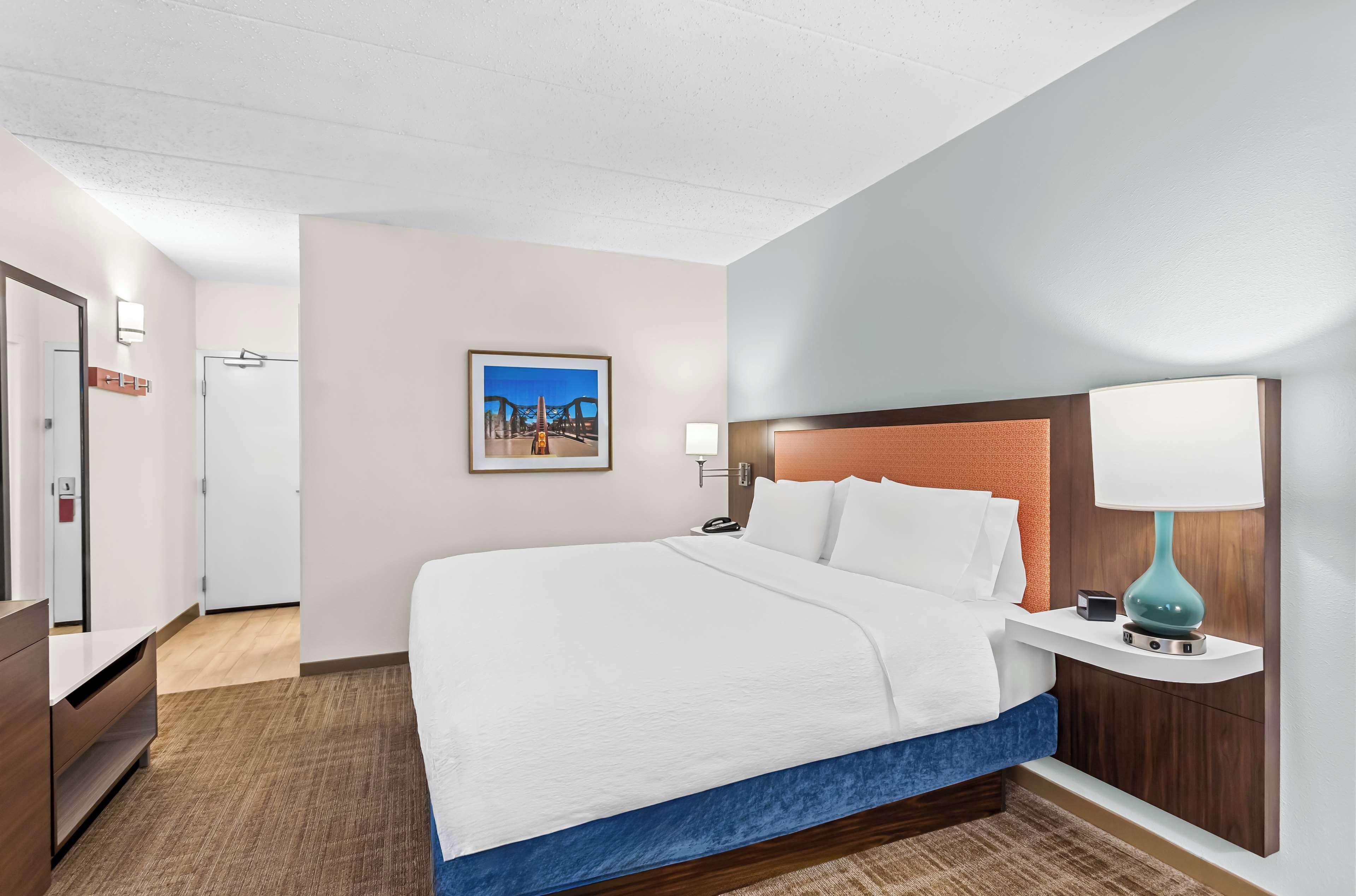 Habitación de hotel con una cama grande, una mesita de noche con lámpara, un cuadro enmarcado en la pared y una puerta que da a otra habitación.