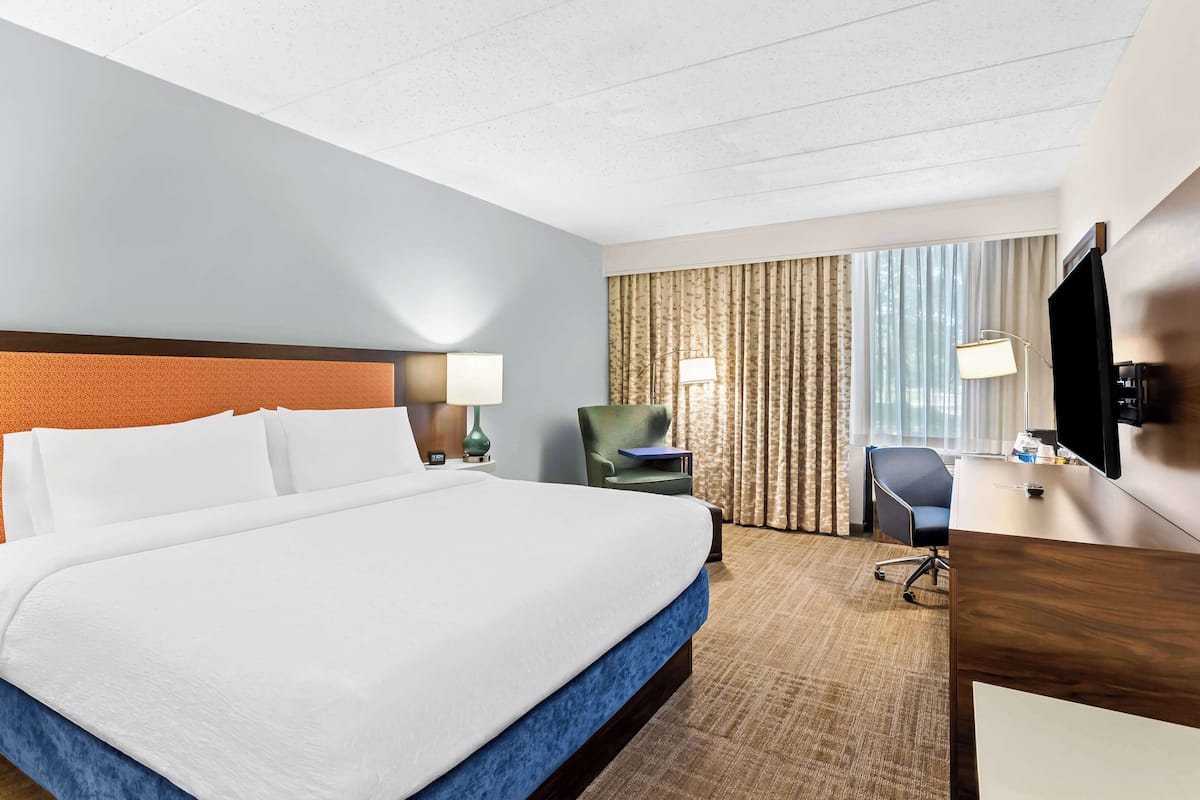 Quarto de hotel com uma cama grande, uma mesa, uma cadeira, uma luminária e uma TV.