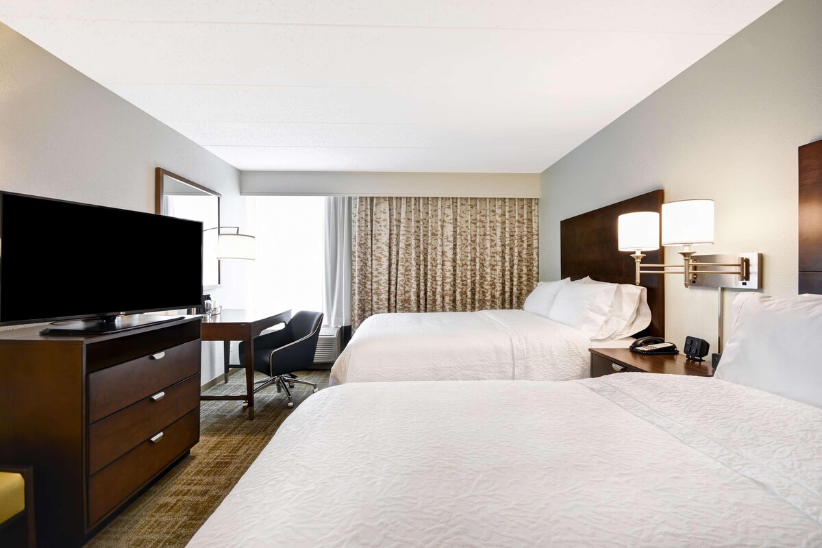 A hotel room with two beds, a TV, a desk with a chair, a lamp, and a window with curtains.