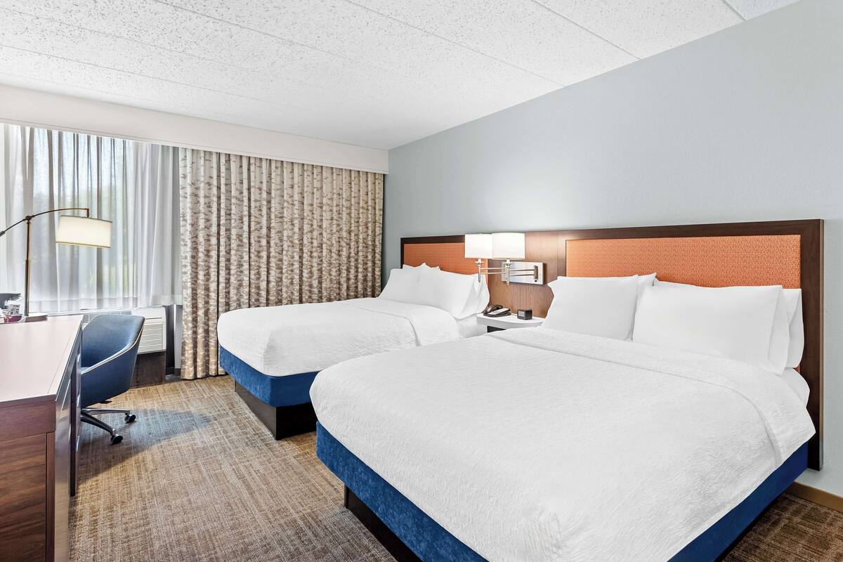 Quarto de hotel com duas camas, uma escrivaninha, uma cadeira e uma janela grande com cortinas.