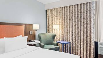 A hotel room with a bed, a green armchair, a small table, a nightstand with a lamp, and a window with patterned curtains.