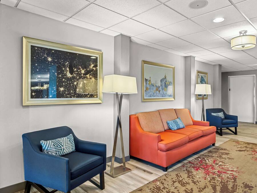 A hotel lobby with a blue armchair, an orange sofa, and a blue armchair with a blue and white pillow. There are two floor lamps and a large painting on the wall.