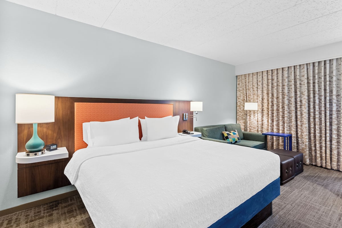 Quarto de hotel com uma cama grande, duas mesas de cabeceira com abajures, uma poltrona verde, uma pequena mesa lateral azul e uma cortina estampada.