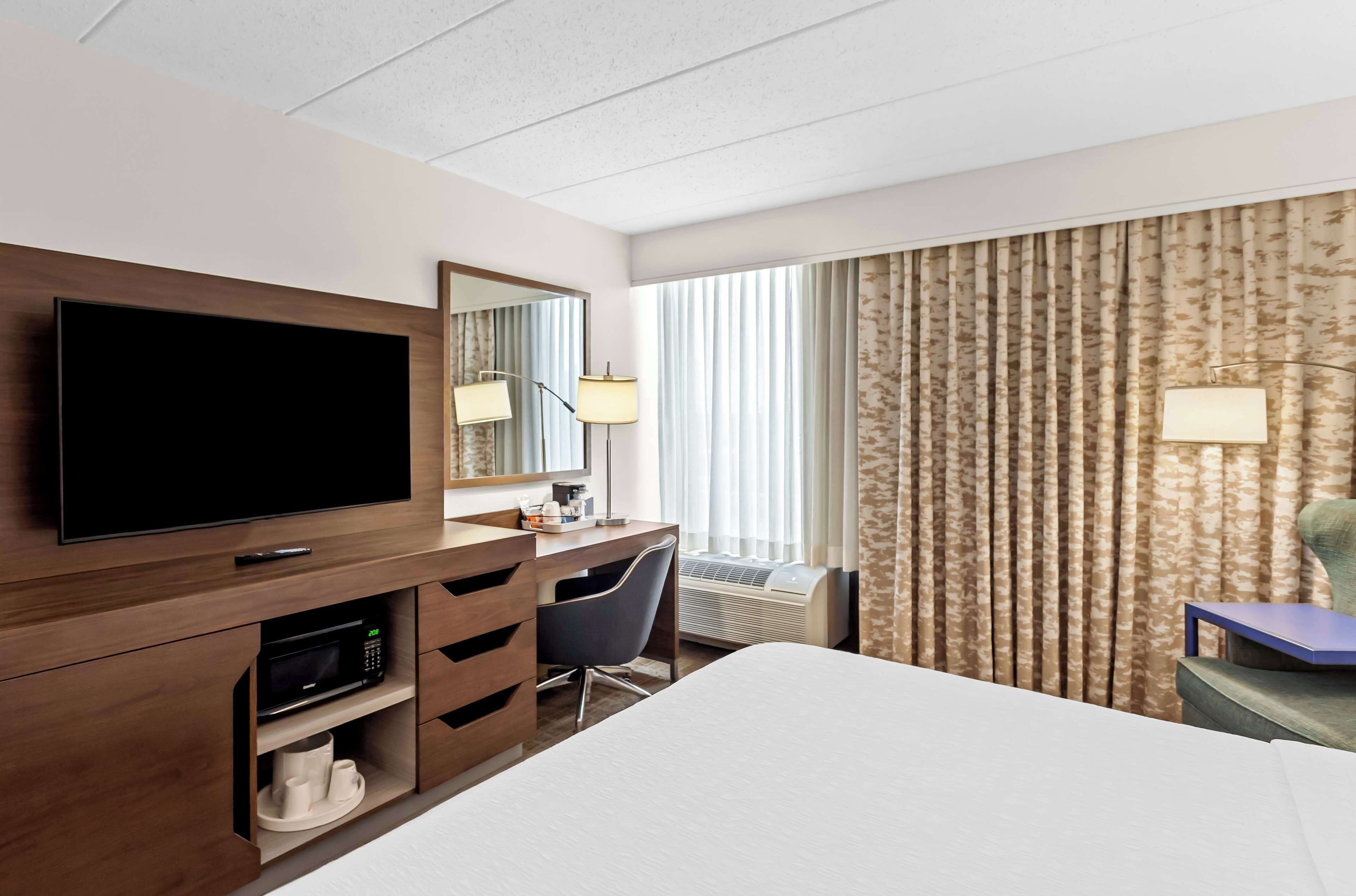 Hotelkamer met flatscreen televisie, bureau met lamp, stoel, bed en groot raam met gordijnen.