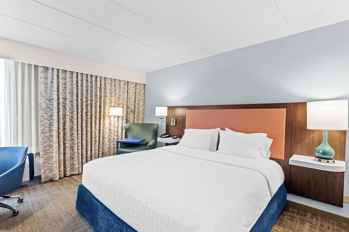 Quarto de hotel com uma cama grande, uma escrivaninha com abajur, uma cadeira e vista para o exterior pela janela.