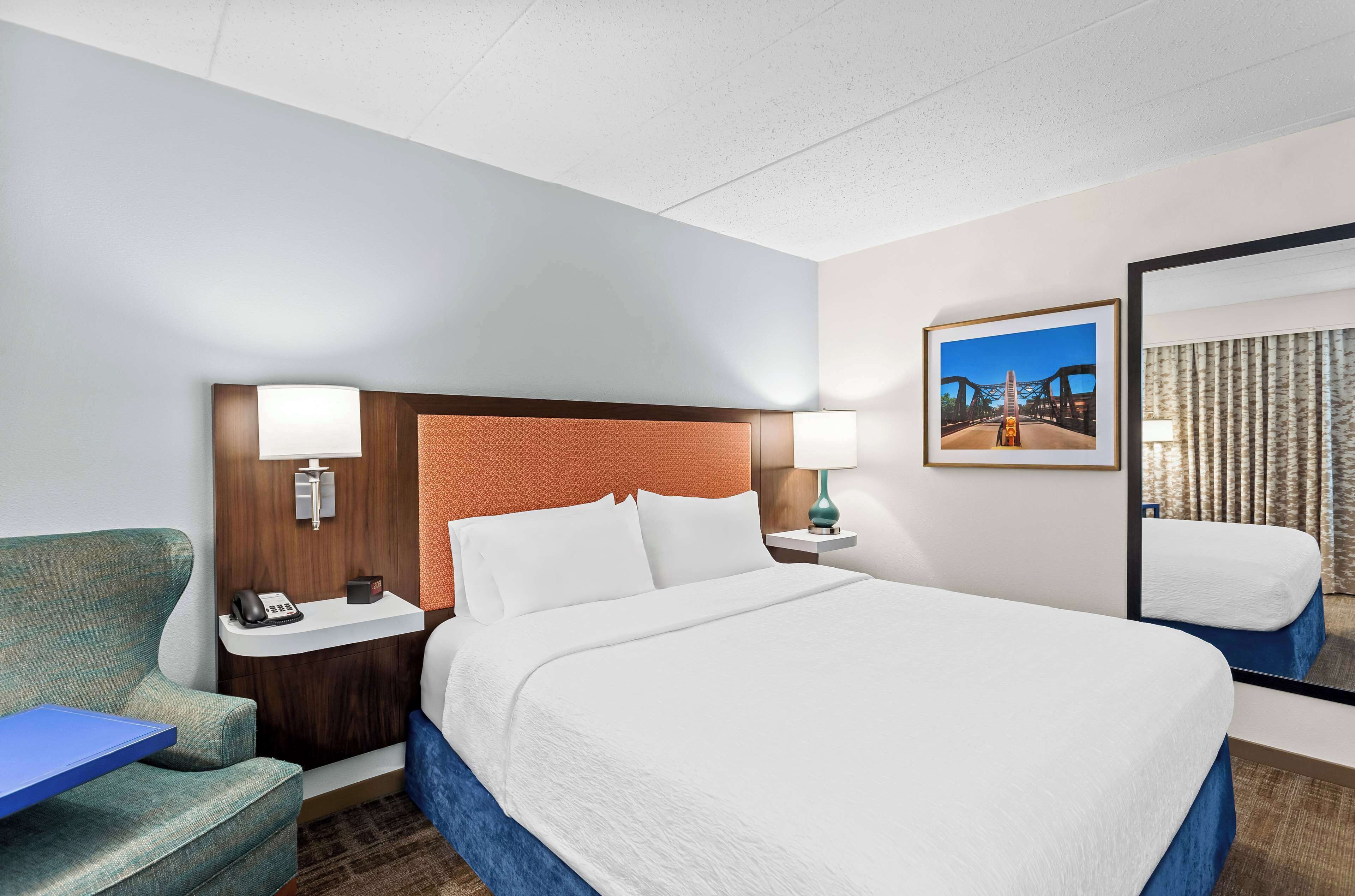A hotel room with a large bed, a desk with a lamp, a chair, and a picture on the wall.