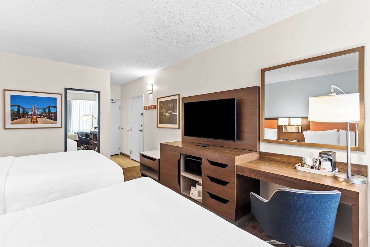 Quarto de hotel com cama, escrivaninha, televisão, cadeira e um espelho grande.