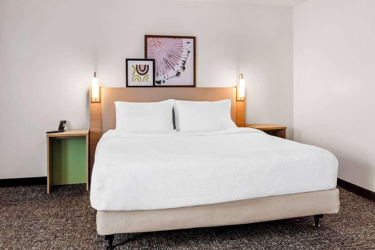 A hotel room with a large bed, two bedside tables, a wall-mounted artwork, and two wall-mounted lamps.