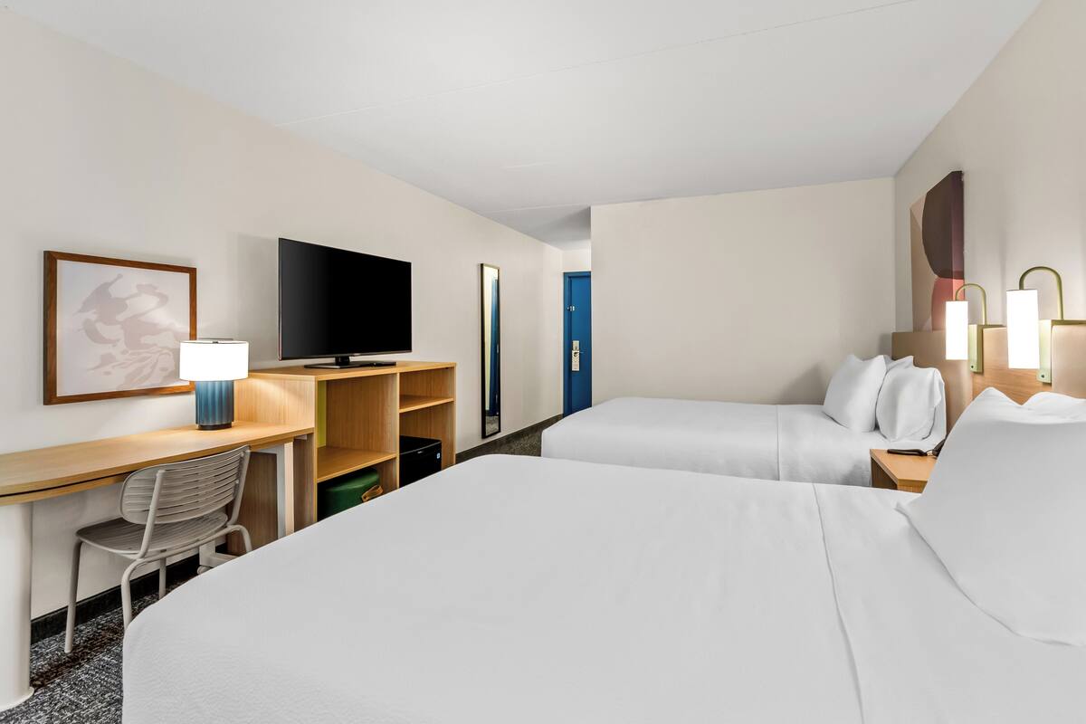 A hotel room with two beds, a desk with a lamp, a television, and a bookshelf.