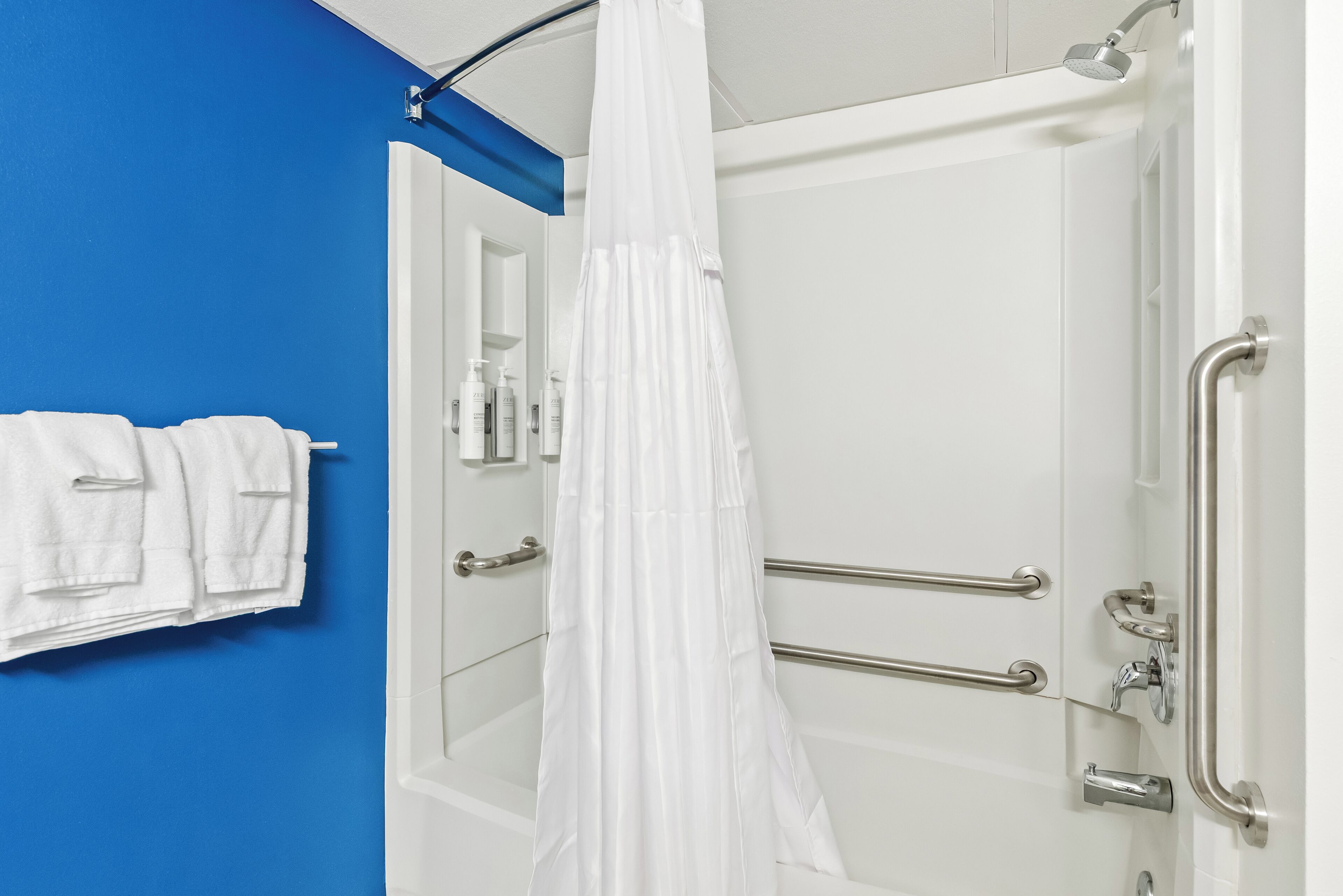 Un baño con pared azul, cortina de ducha blanca y barras de apoyo.