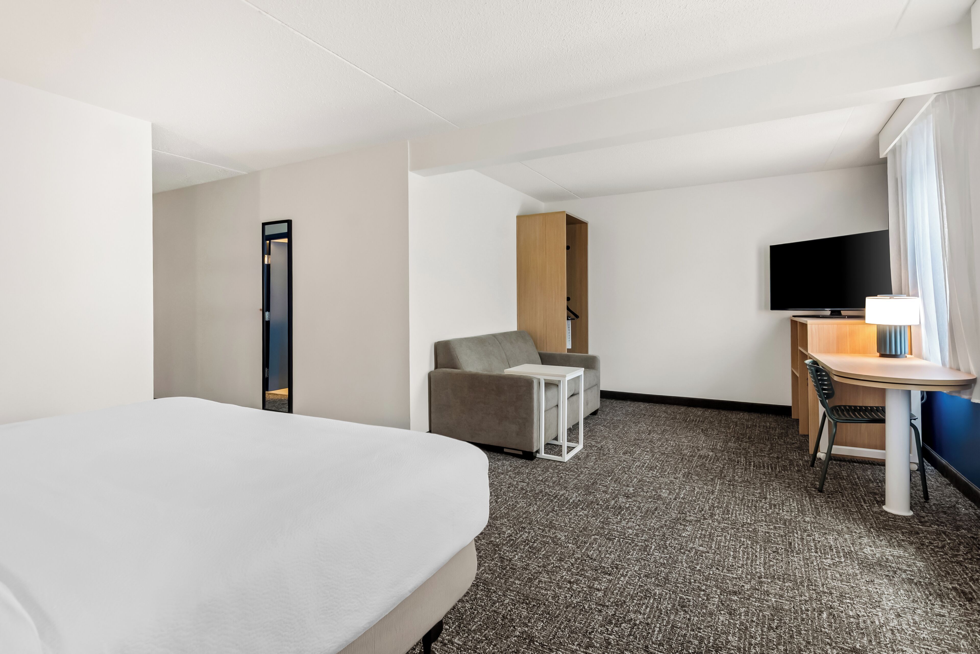 A hotel room with a bed, a desk with a chair, a sofa, a TV, and a window with curtains.