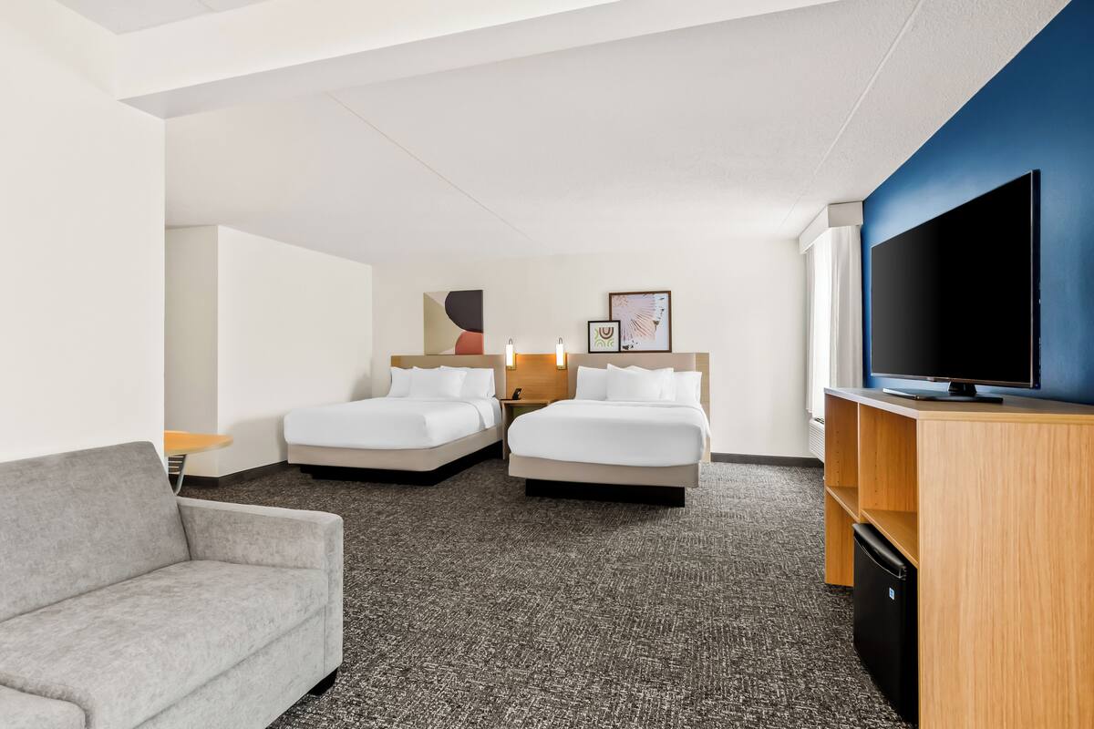 A hotel room with two beds, a gray sofa, a TV, and a desk with a computer.