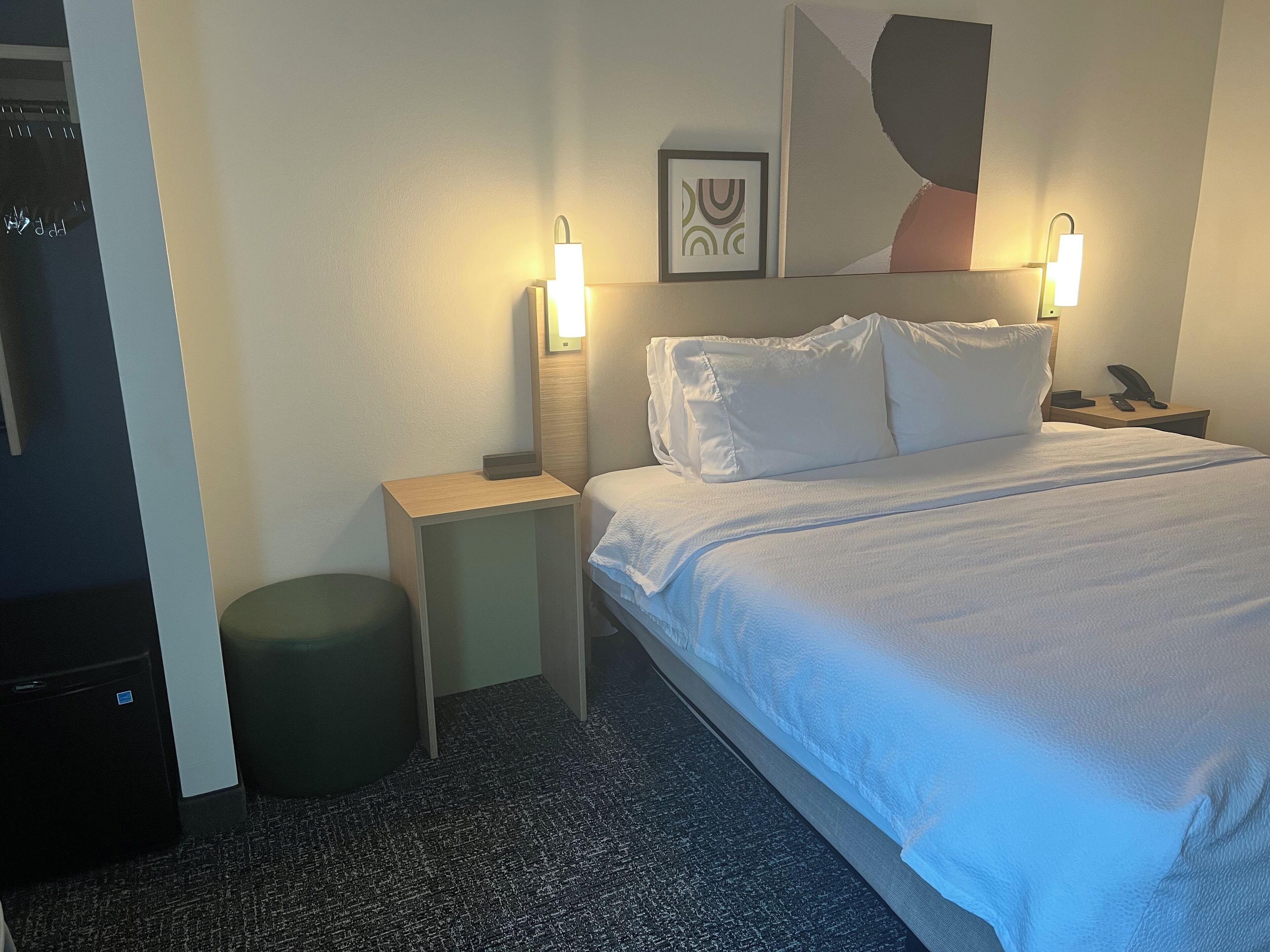 A hotel room with a bed, two bedside lamps, a nightstand, a small stool, and a framed picture on the wall.