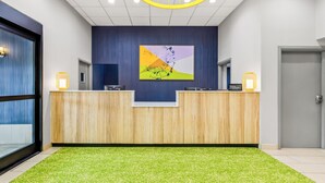 Un espace d’accueil d’hôtel avec un comptoir de réception en bois, une peinture abstraite aux couleurs vives et une moquette verte.