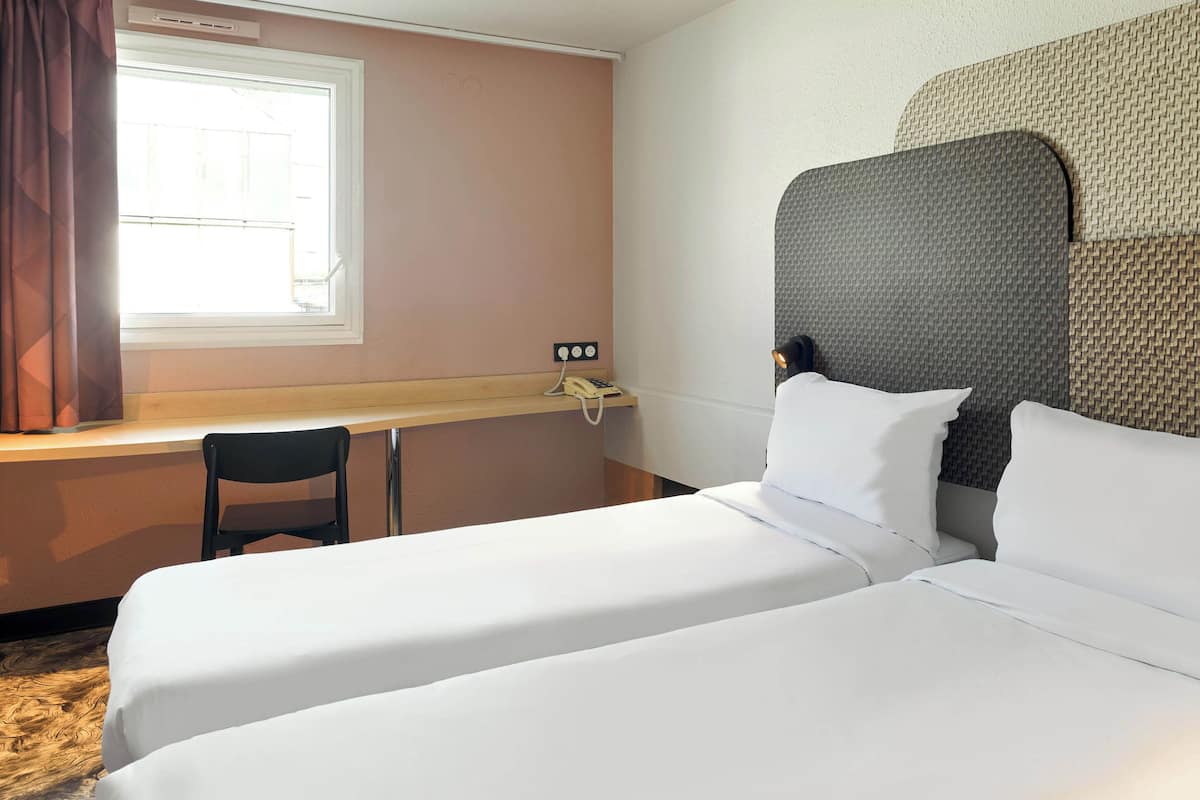 A hotel room with two single beds, a desk with a chair, a telephone, and a window with curtains.
