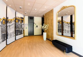 A hallway with a wooden floor, a black bench, a large mirror, and a white vase with flowers.