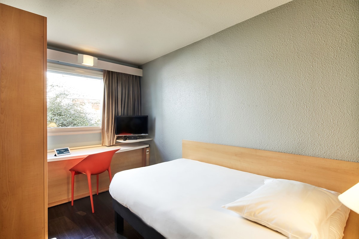 A hotel room with a bed, a desk with a chair, a television, and a window with curtains.