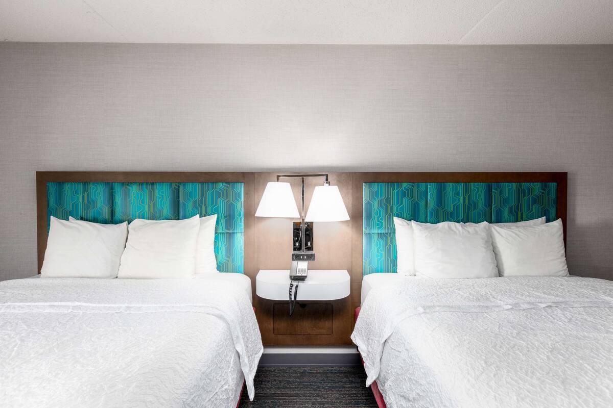 A hotel room with two beds, a wooden headboard, a built-in phone, and teal patterned curtains.