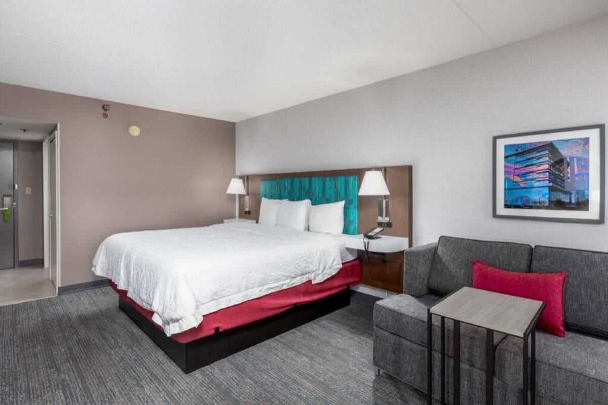 A hotel room with a bed, a sofa, a small table, a nightstand, and a painting on the wall.