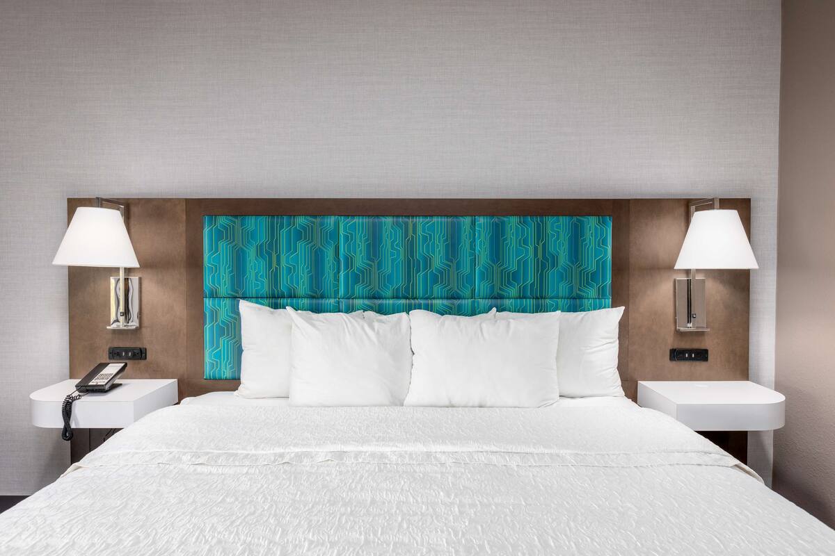 A hotel room with a large bed, white bedding, a teal patterned headboard, two bedside tables with lamps, and a phone on one table.