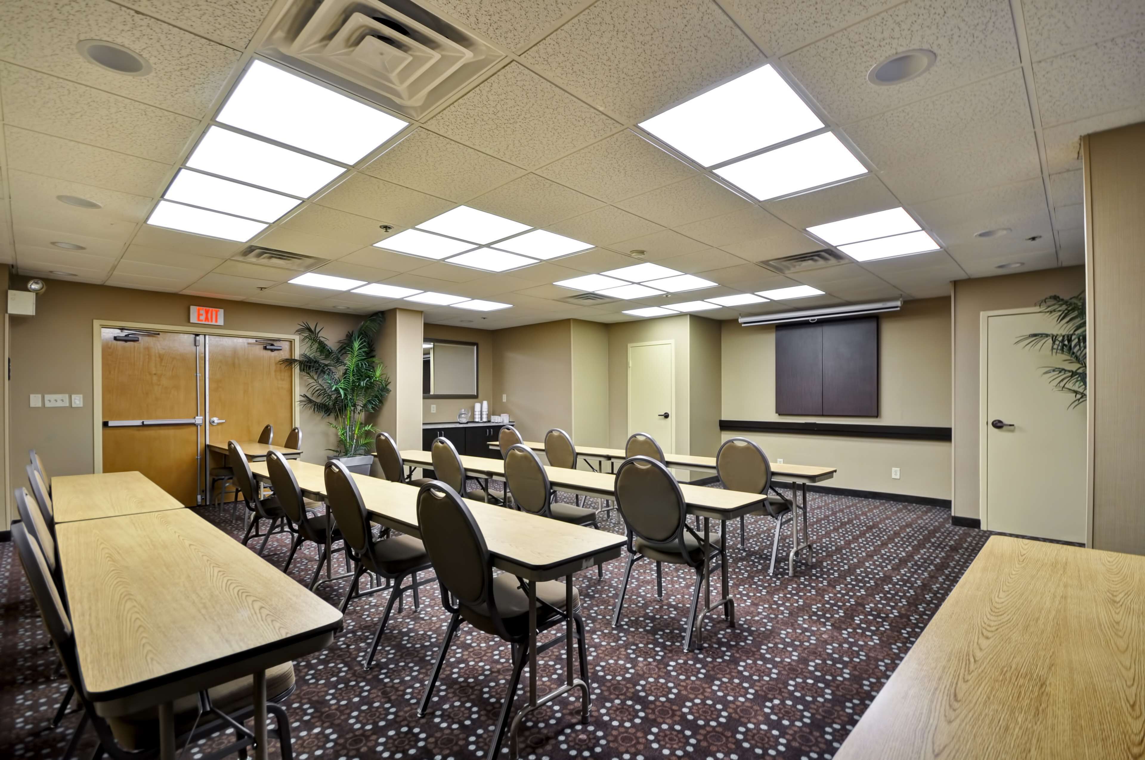 A conference room with multiple tables and chairs, a projector screen, and a door with an exit sign.