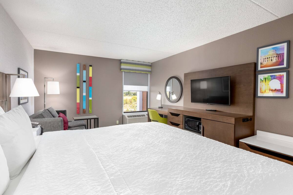 A modern hotel room with a large bed, a desk, a television, and a window with a view of greenery.
