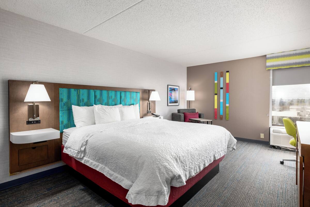 A hotel room with a large bed, bedside lamps, a small table, a chair, a window, and a wall-mounted air conditioning unit.