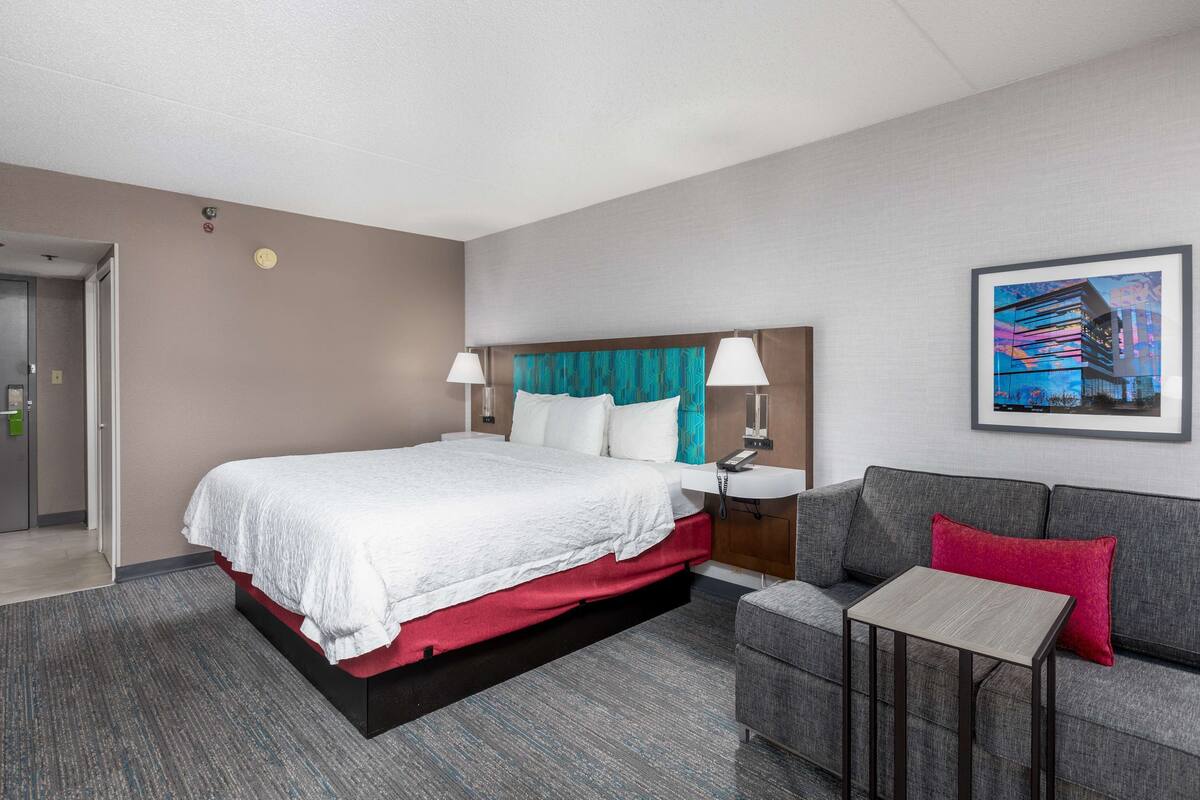 A hotel room with a large bed, a gray sofa, a small table, and a painting on the wall.