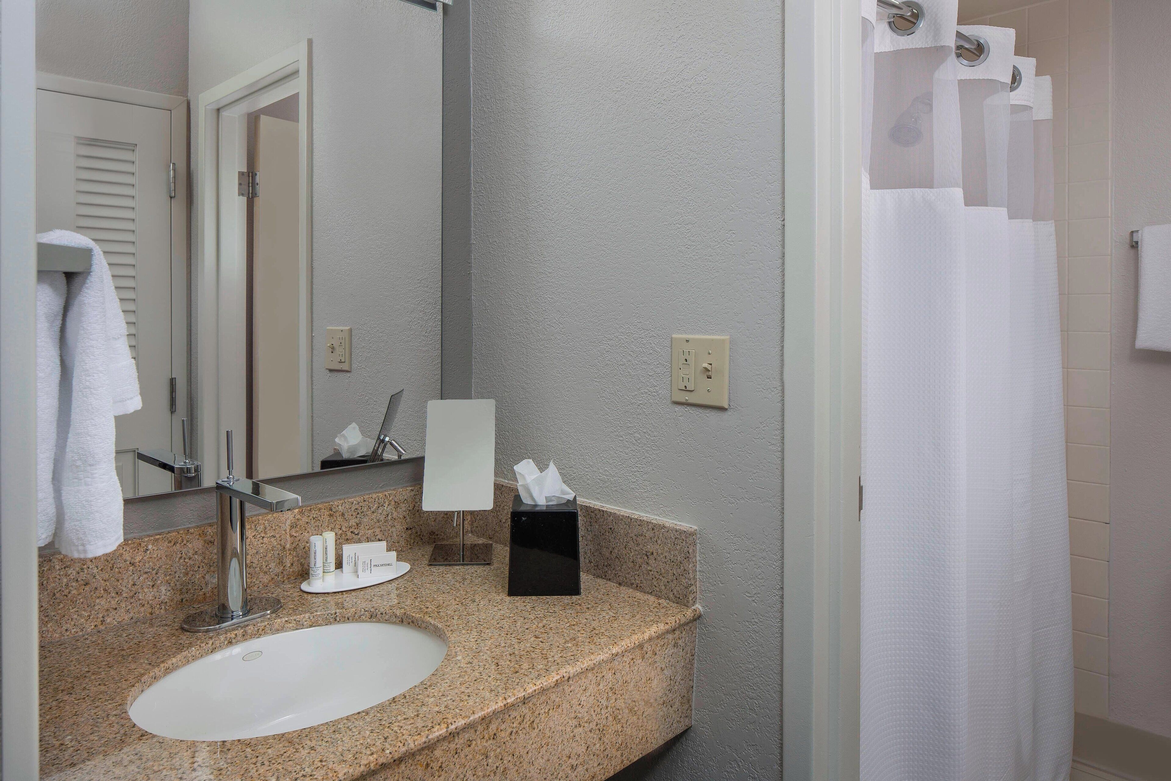 A bathroom with a granite countertop, a white sink, a mirror, a towel rack, a tissue box, and a shower curtain.