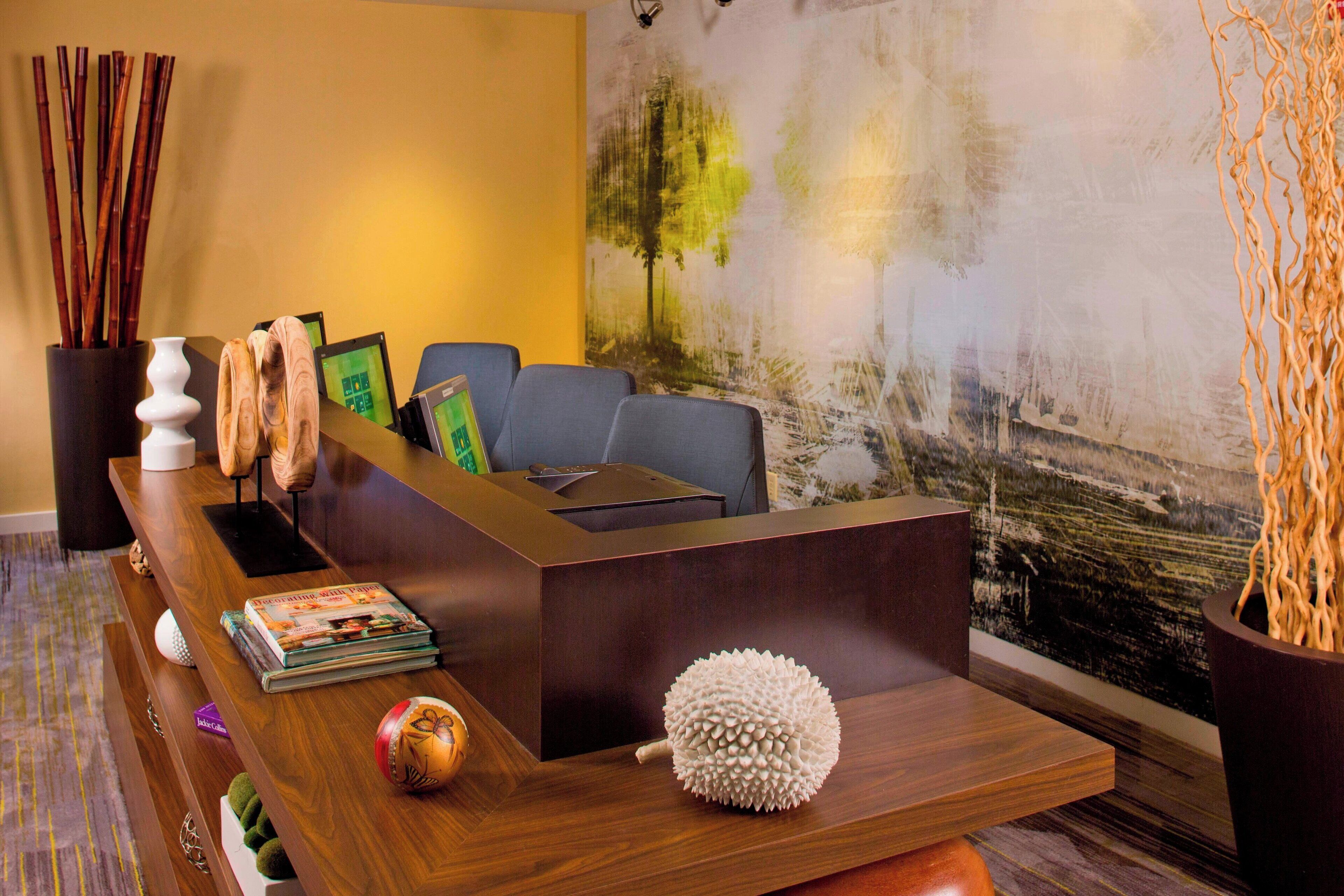 Een receptiebalie met een modern ontwerp, afgewerkt met hout en voorzien van een flatscreen televisie. Op de balie staan diverse decoratieve objecten, waaronder een bolvormige sculptuur en een vaas met een plant. Op de achtergrond hangt een groot abstract schilderij en een wandlamp.