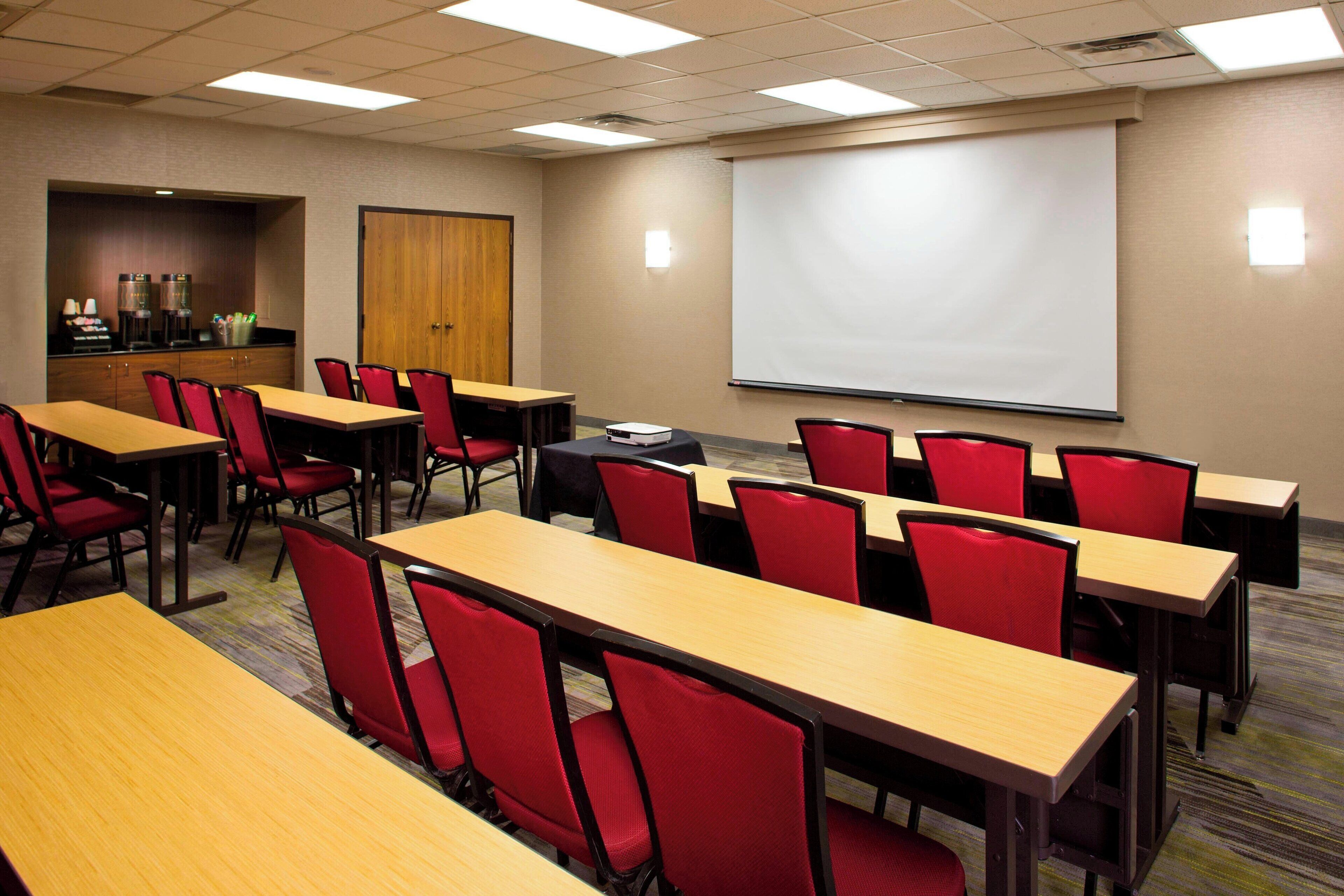 Une salle de conférence équipée de plusieurs tables et de chaises rouges, d’un écran de projection et d’un coin café.
