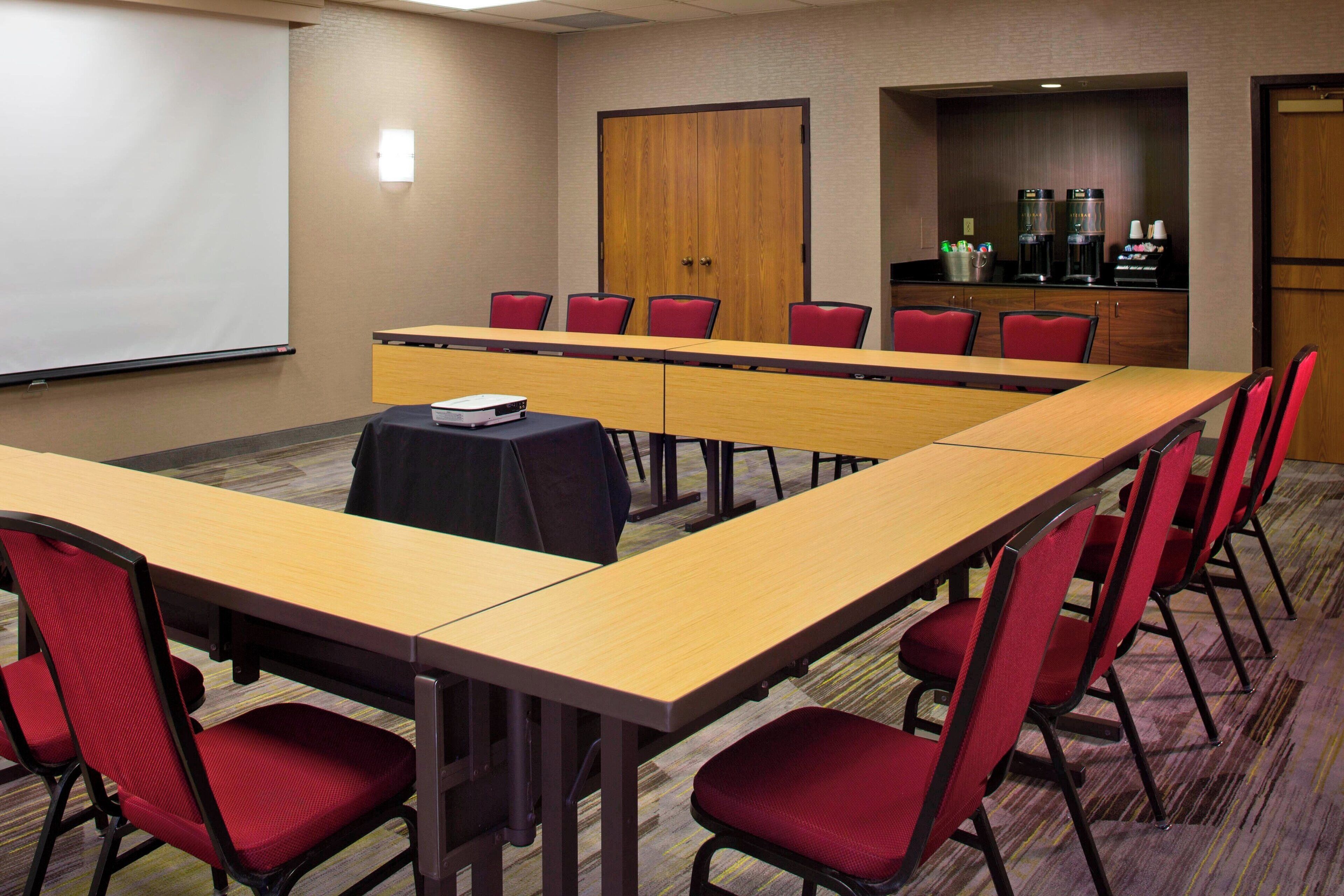 A conference room with a long table, red chairs, a projector screen, and a coffee station.