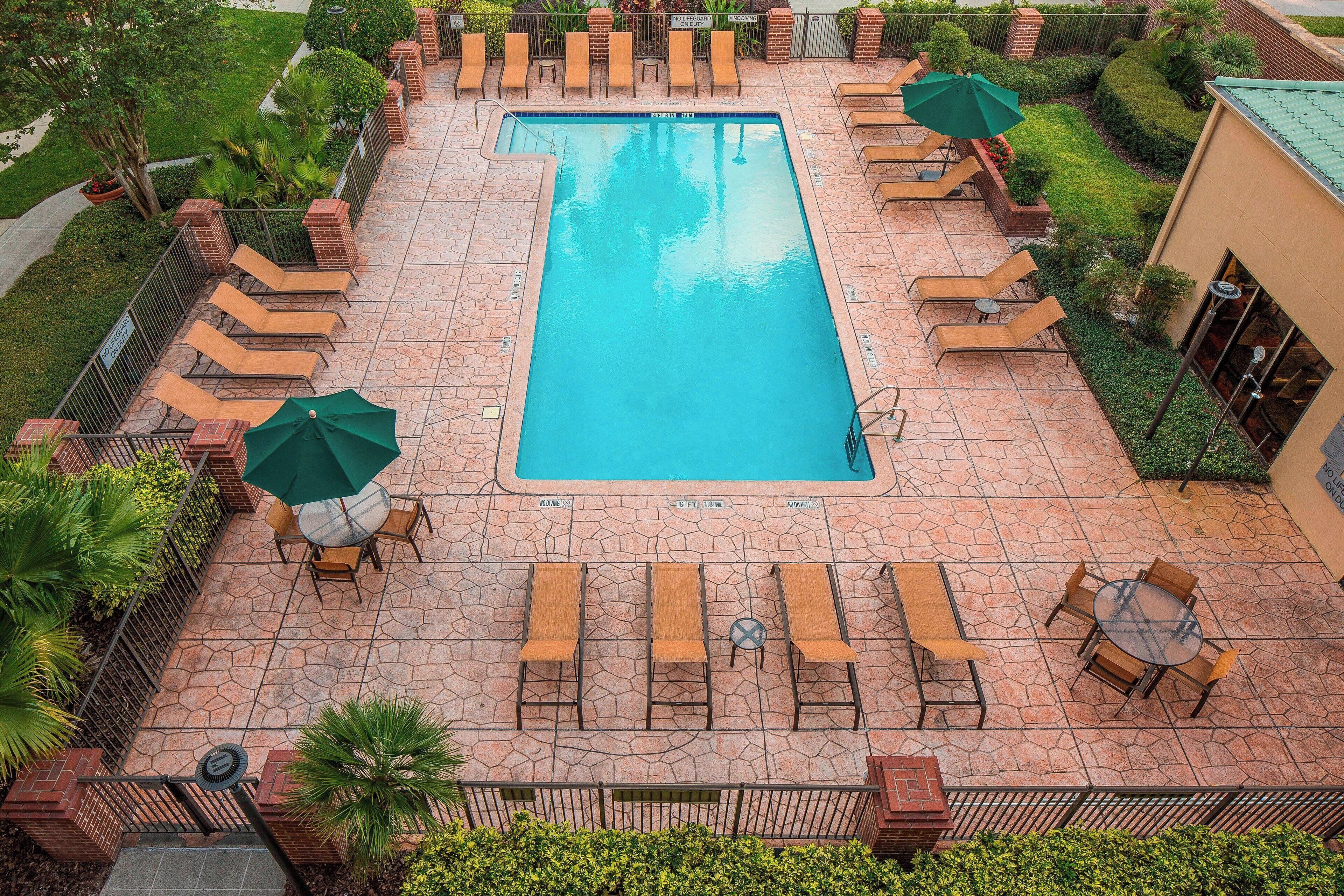 An aerial view of a rectangular swimming pool surrounded by lounge chairs and umbrellas, with a stone patio and greenery.