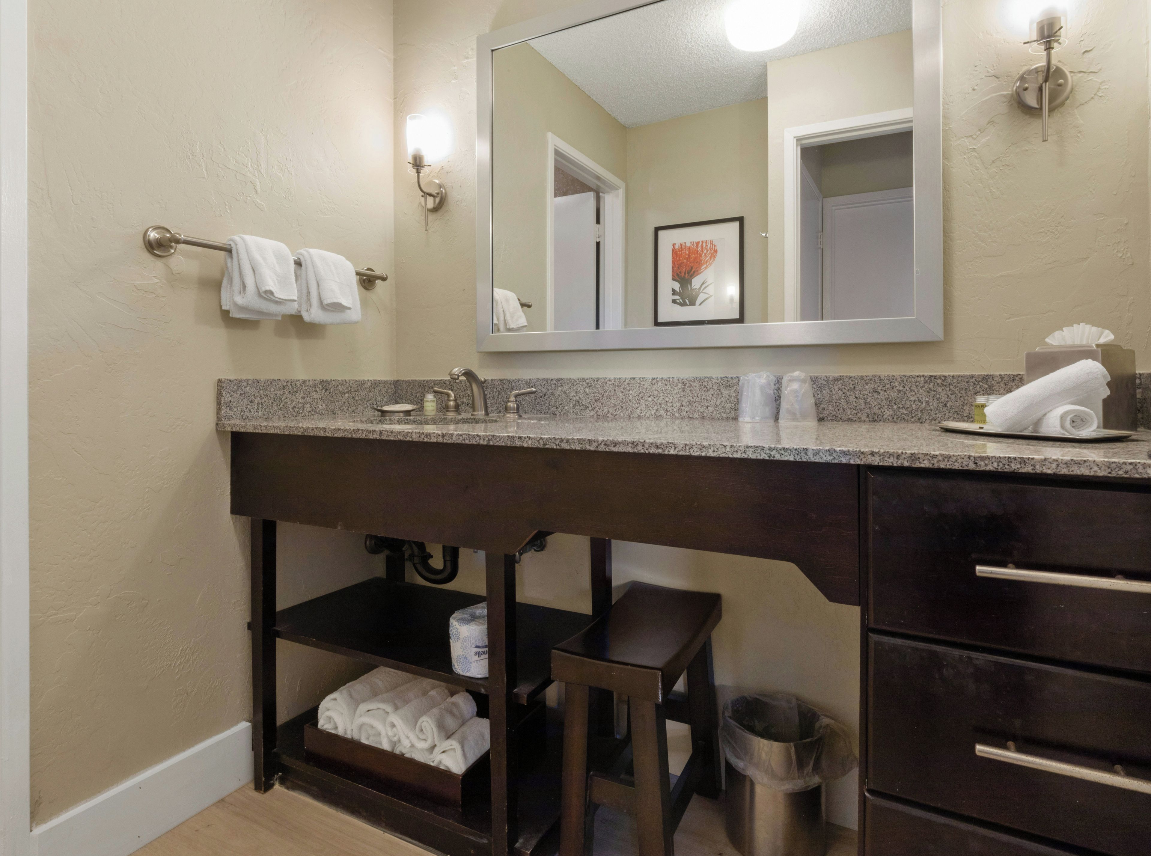 A bathroom with a granite countertop, a sink, a mirror, a towel rack, a trash can, and a shelf with towels.