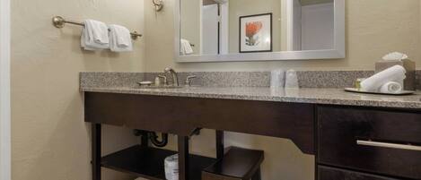 A bathroom with a granite countertop, a sink, a mirror, a towel rack, a trash can, and a shelf with towels.