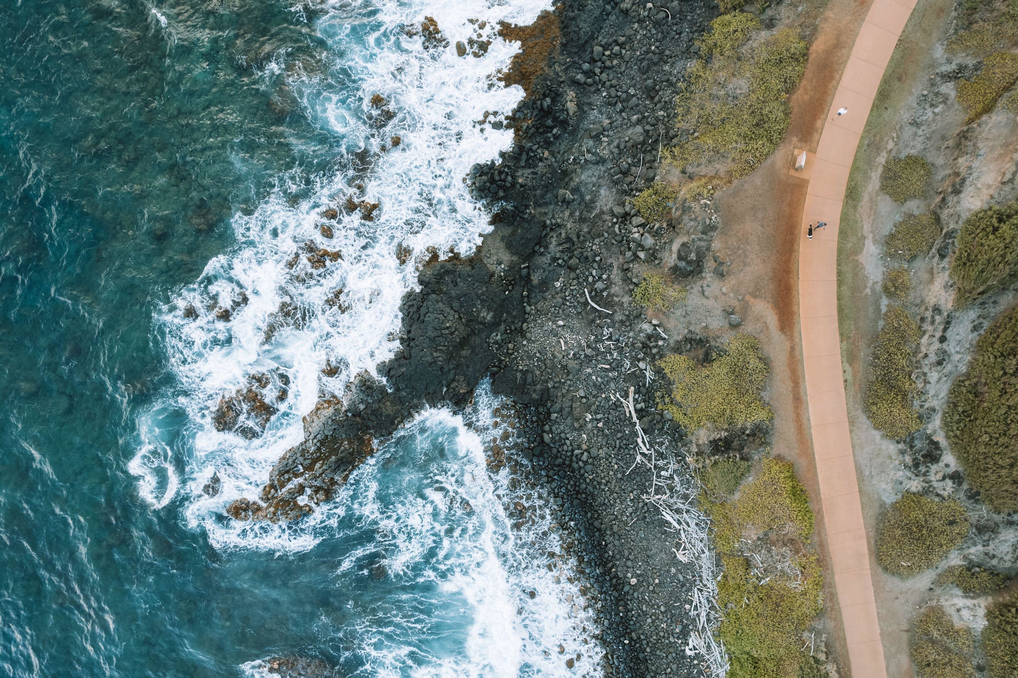 Vista aérea de una zona costera con un paseo pavimentado a lo largo de la orilla, terreno rocoso y olas rompiendo en la playa.