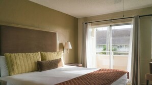 A hotel room with a large bed, a nightstand, a lamp, and a view of the outside through a window.