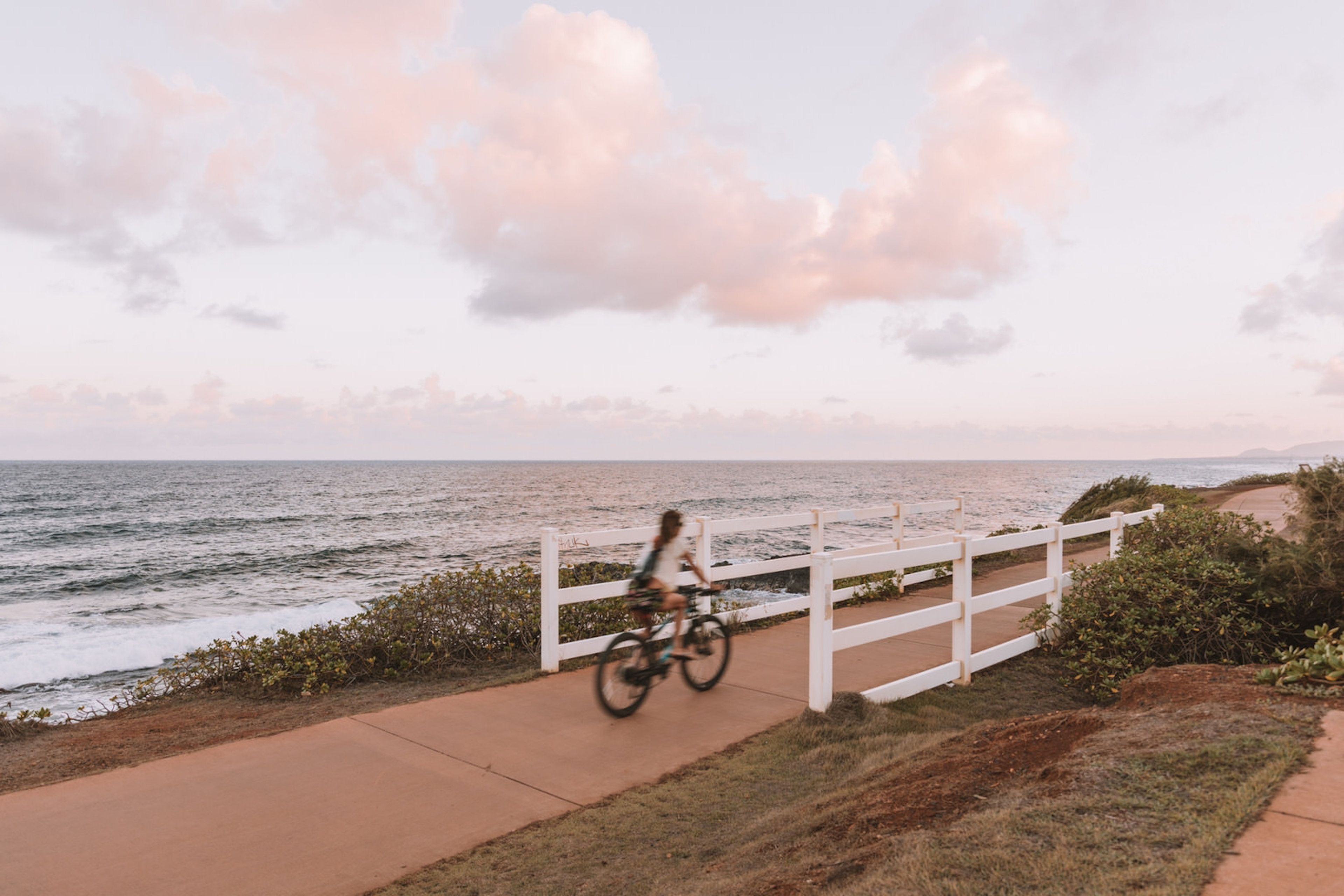 A coastal walkway with a white fence, a person riding a bicycle, and the ocean in the background.
