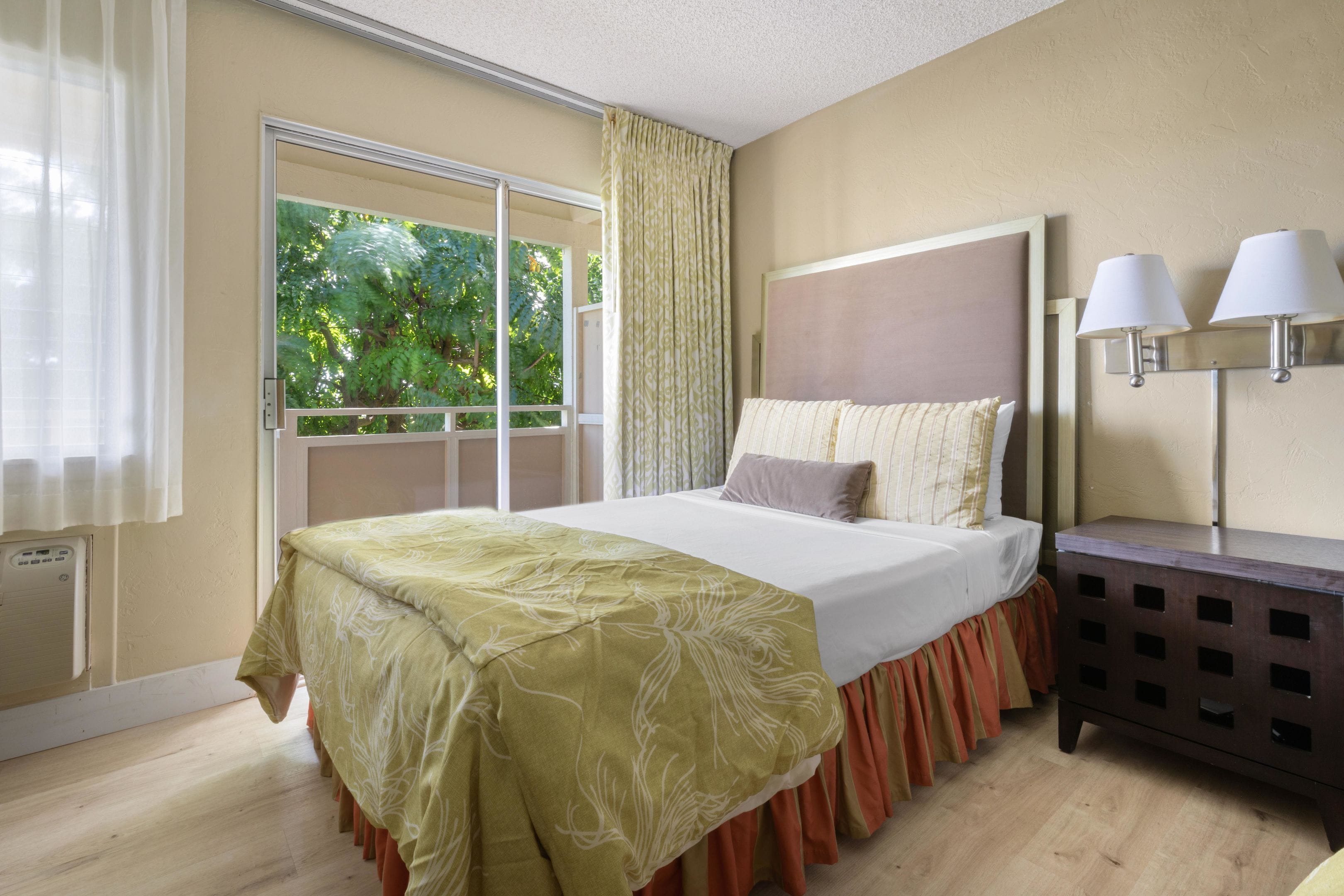 A hotel room with a bed, a nightstand, a window with curtains, and a view of greenery outside.