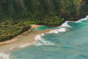 An aerial view of a coastal area with a beach, clear turquoise waters, and dense green forested hills. - Plantation Hale Suites (Kapaa)