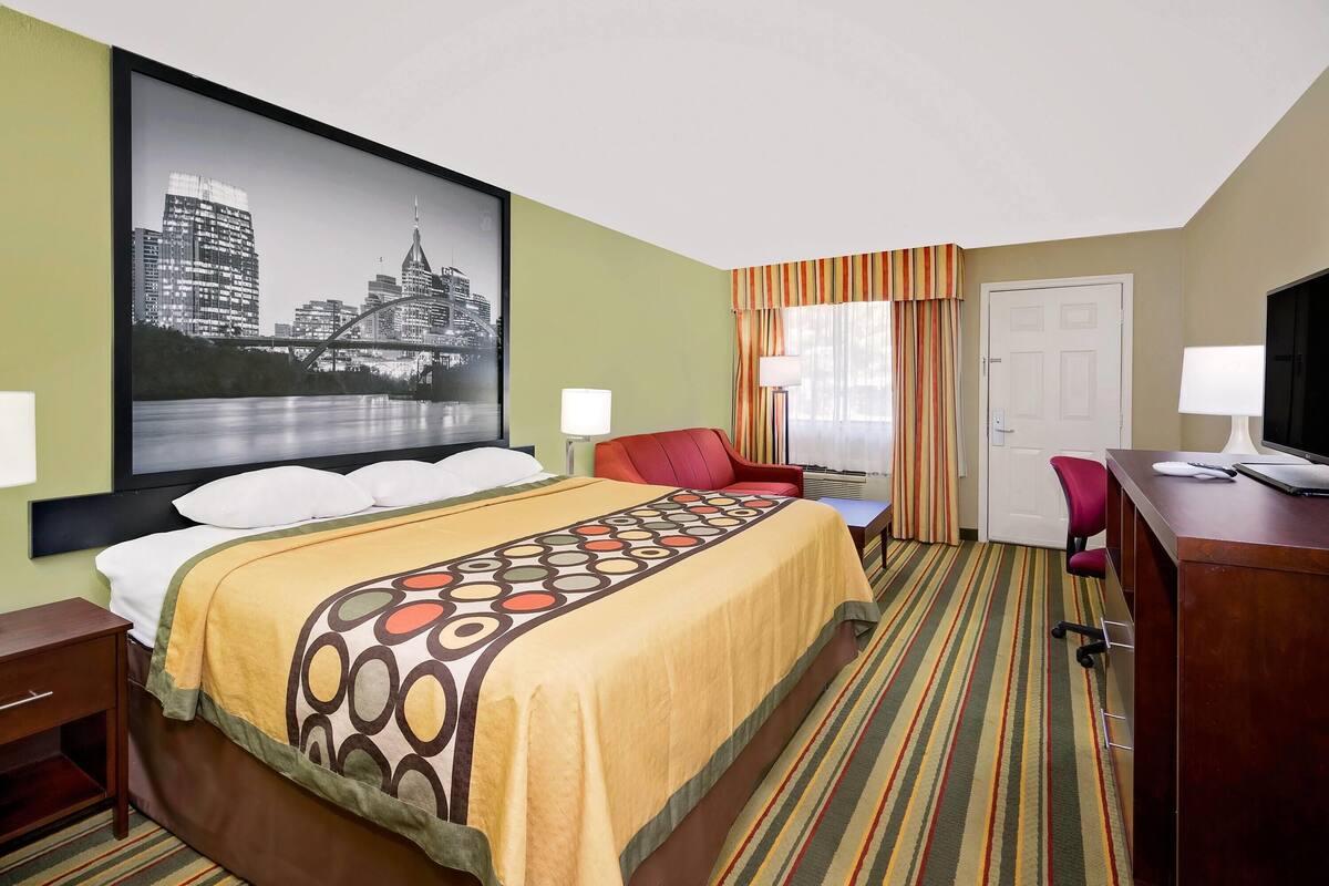 A hotel room with a bed, a desk, a chair, a TV, and a large cityscape painting on the wall.