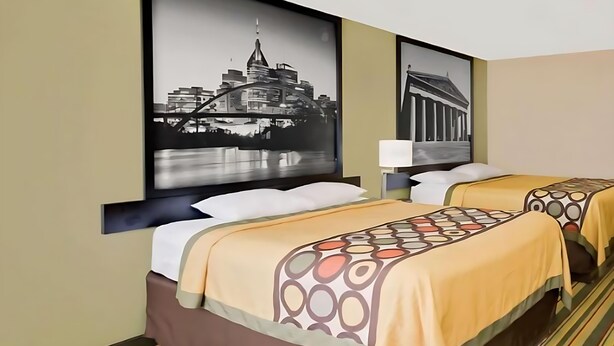 A hotel room with two beds, each with a patterned bedspread, a nightstand with a lamp, and two large framed pictures on the wall. - Super 8 by Wyndham Hermitage Nashville (Hermitage)